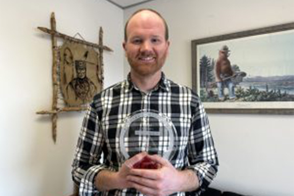 A smiling man with a beard, wearing a black and white plaid shirt, stands indoors holding a clear circular award.