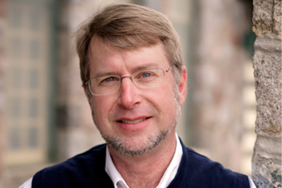 A man with short light brown hair wearing glasses smiles.