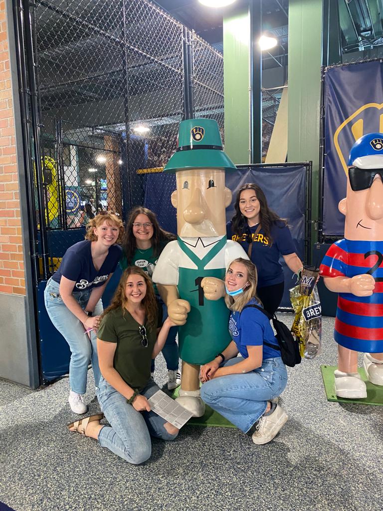 A photo featuring residents of Stockwerk Deutsch posing with a statue of a man in traditional German attire. The statue has a hat that features the Milwaukee Brewers logo.