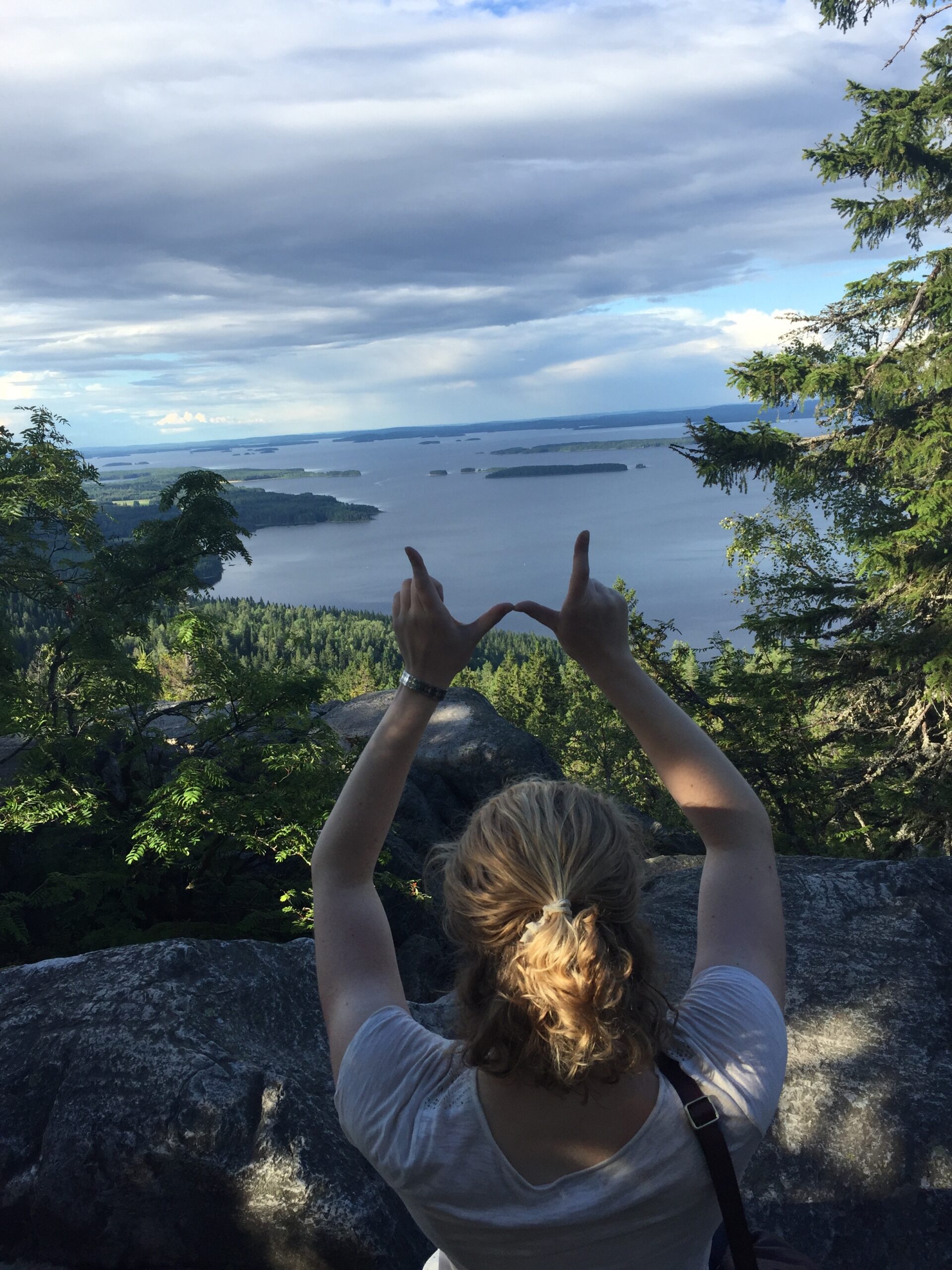 A grad student holds their hands up in a W formation over a lake at the Koli National Park in Finland. 